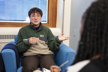 College student talking and gesturing with their hands while sitting across from a person who is writing on a clipboard