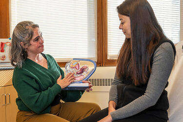 a clinicial and pateint sit facing each other in an exam room. The clinician holds a model of the female reproductive system