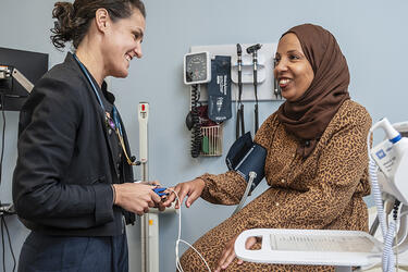 A clinician stands facing a seated patient while placing a pulse oximeter on the patient’s finger