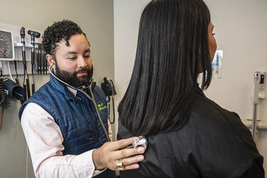 A clinician stands next to a patient and holds a stethoscope bell against their back