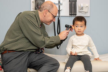 clinical sits on a treatment table next to a toddler while examining the child’s inner ear with an otoscope 