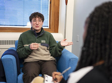 College student talking and gesturing with their hands while sitting across from a person who is writing on a clipboard