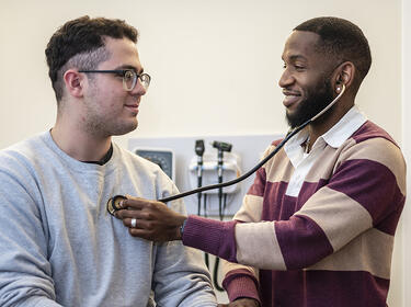 Clinician faces a patient while listening to their heart with a stethoscope