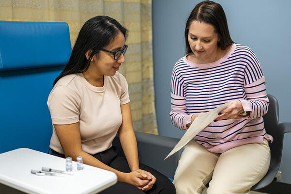 A clinician sits next to a patient while showing the patient a document. Two syringes and medication vials sit on a small table beside the patient 