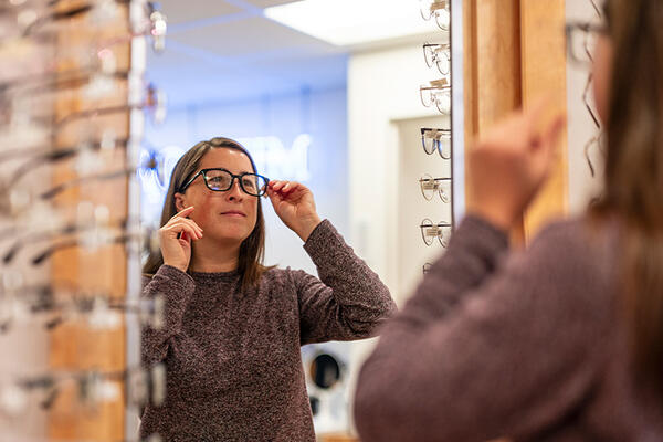 reflection in a mirror of a person trying on eyeglasses at the MIT Health optical shop. Racks of eye glasses are on both sides of the mirror